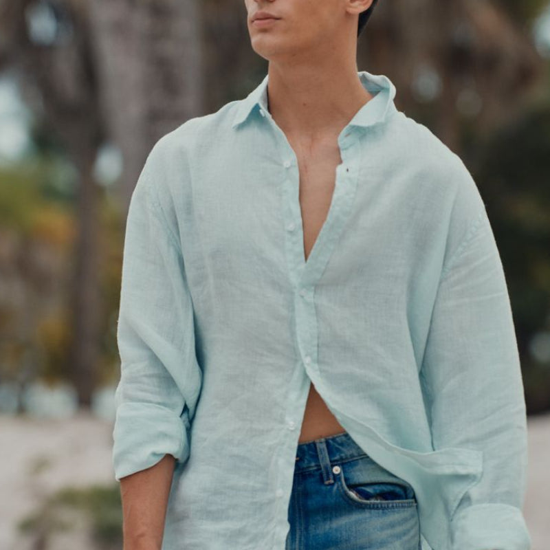 Man wearing a light blue shirt and jeans standing outdoors with trees in the background