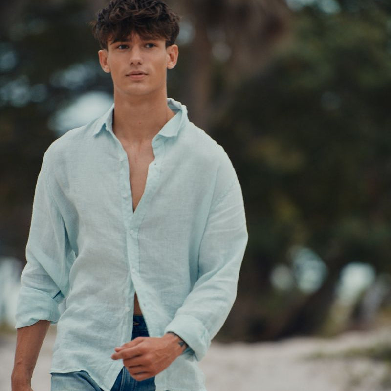 Man wearing a light blue shirt and jeans standing on a beach with trees in the background