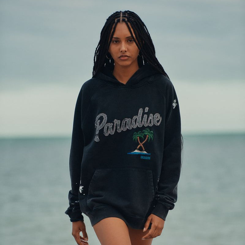 Woman wearing a black okaicos hoodie with 'Paradise' text and palm tree design on a beach.