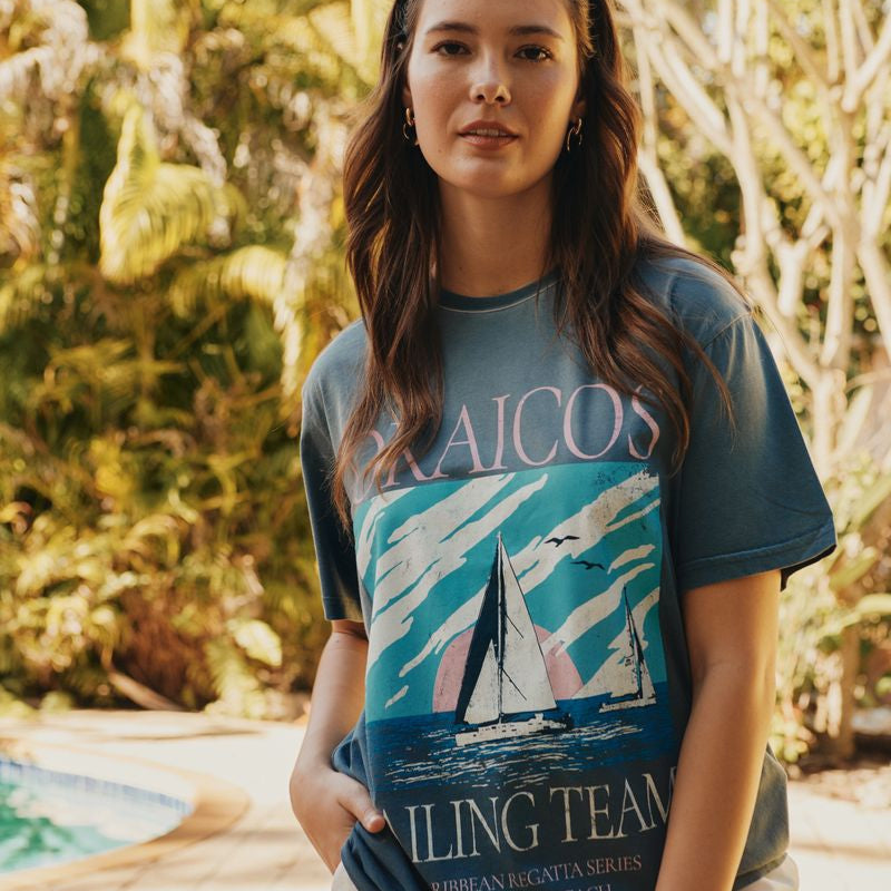 Woman wearing an Okaicos t-shirt with a sailboat design and text outdoors.