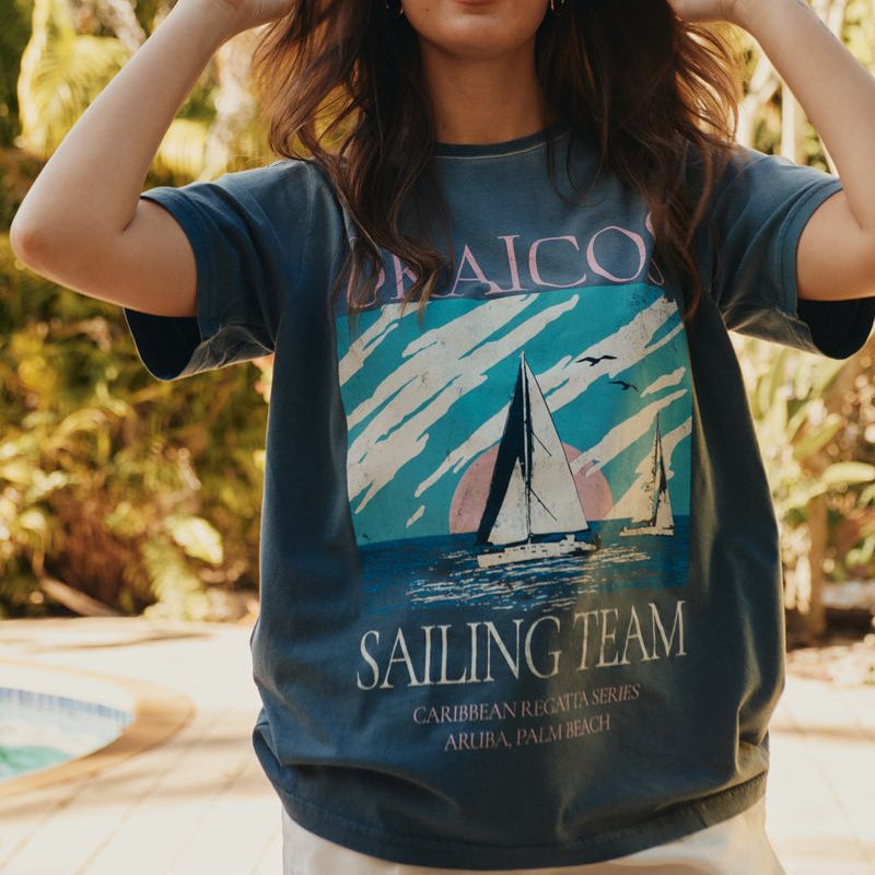 Woman wearing a blue Okaicos t-shirt with a sailboat design and text outdoors.