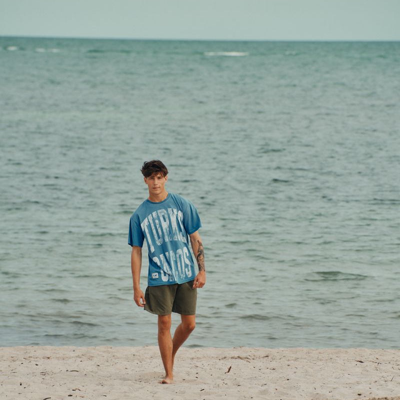Person walking on a beach with okaicos blue turks & caicos print t-shirt with the ocean in the background
