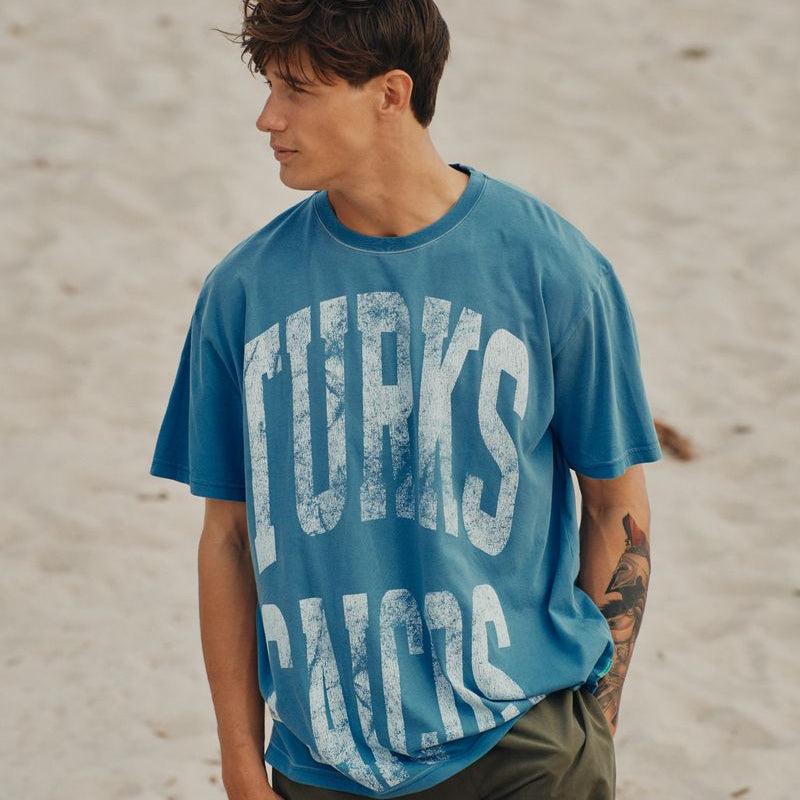 Man wearing a blue okaicos t-shirt with 'Turks & Caicos' text on a sandy background
