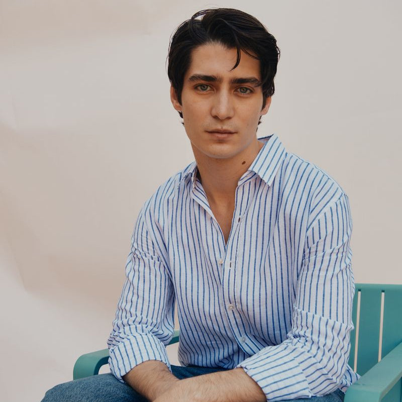 Man wearing a blue striped okaicos shirt and jeans sitting on a green chair against a white background