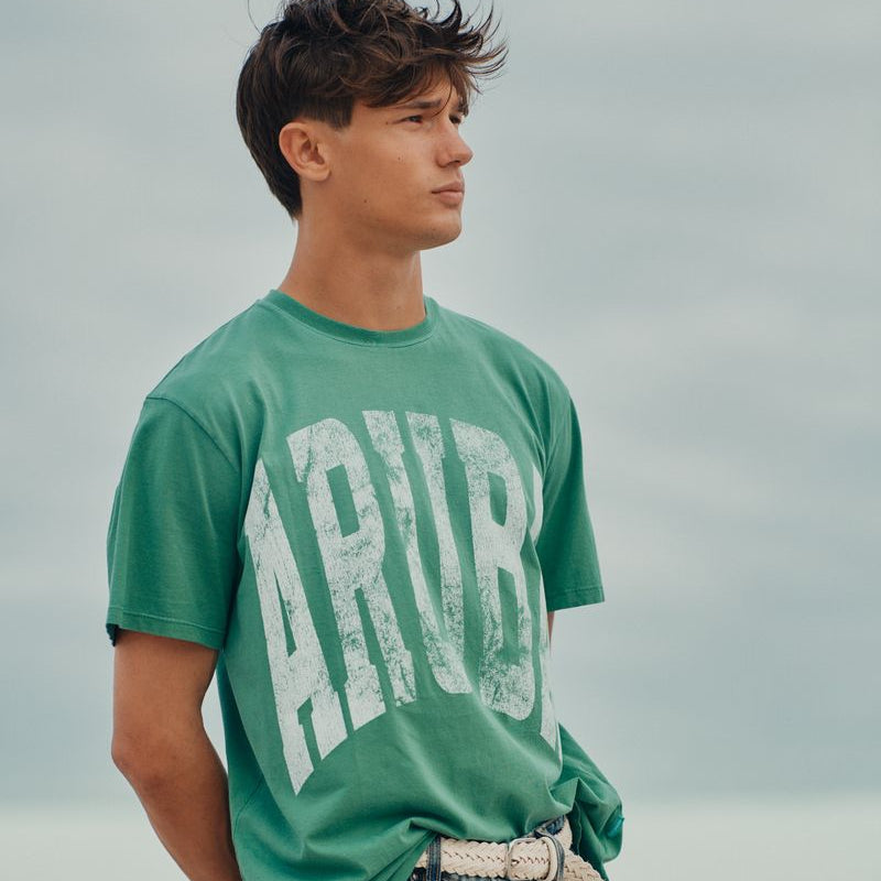 Person wearing a green Okaicos 'Aruba' t-shirt standing against a blurred natural background