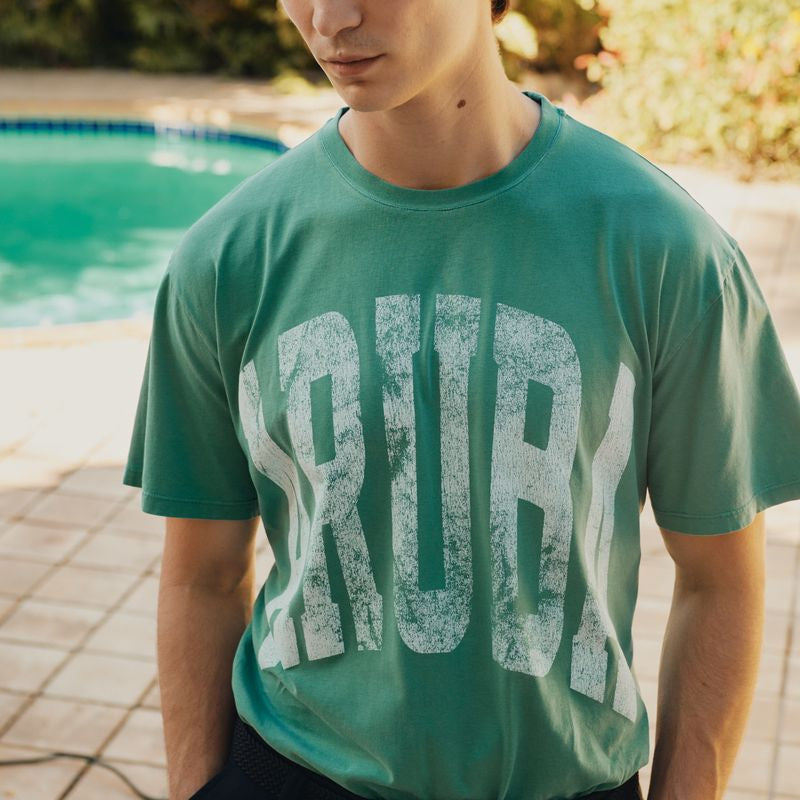 Person wearing a green Okaicos t-shirt with 'CRUE' printed on it, standing by a pool.