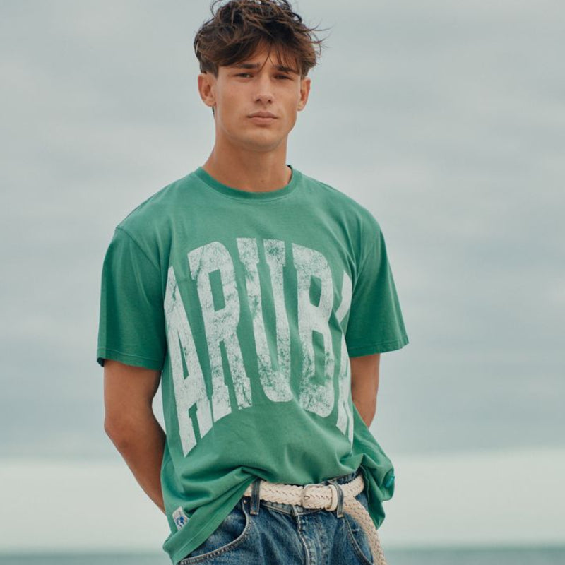 Person wearing a green Okaicos t-shirt with 'ARUBA' text standing by the ocean.