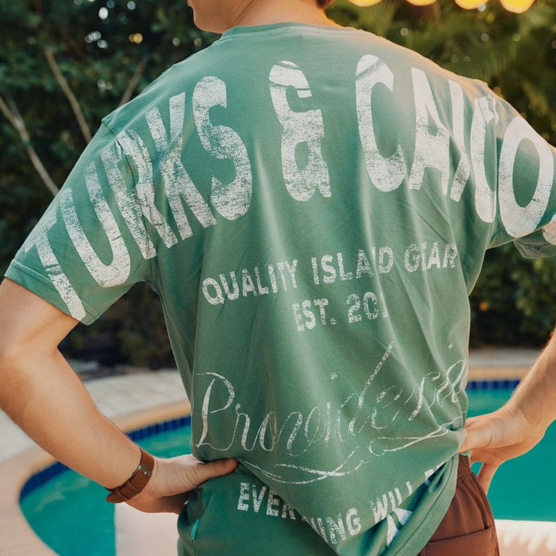 Person wearing a green Okaicos 'Turks & Caicos' t-shirt by a poolside with a yellow umbrella.