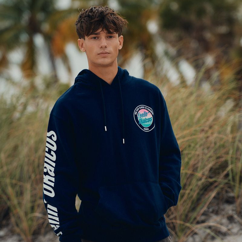 Person wearing a navy blue okaicos hoodie with a logo and text, standing on a beach with palm trees in the background.