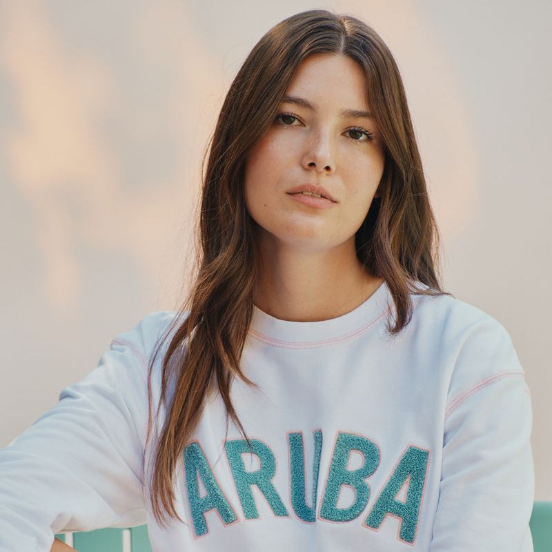 Woman wearing a white okaicos sweatshirt with 'ARUBA' printed on it, sitting against a blurred background.