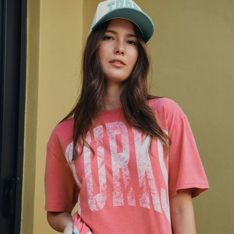 Woman wearing a red pink oversized Okaicos t-shirt with Turks & Caicos text and a cap against a yellow wall.