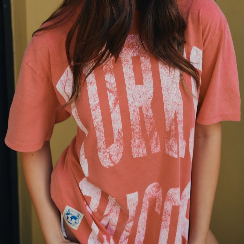 Woman wearing a red coral t-shirt with text and a cap with 'TurKS' on it against a yellow background