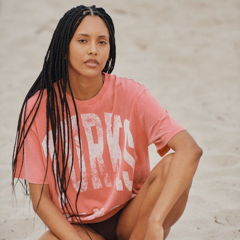 Woman sitting on the sand wearing a red pink Okaicos 'Turks' t-shirt.