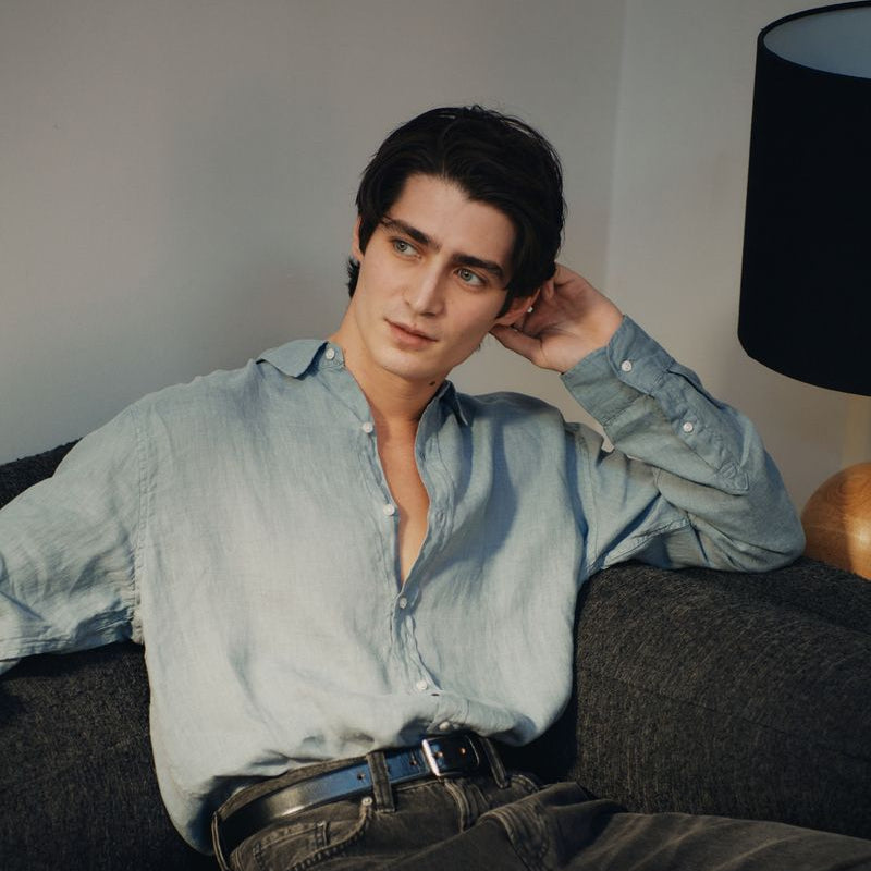 Man sitting on a couch wearing a light grey okaicos shirt and dark jeans