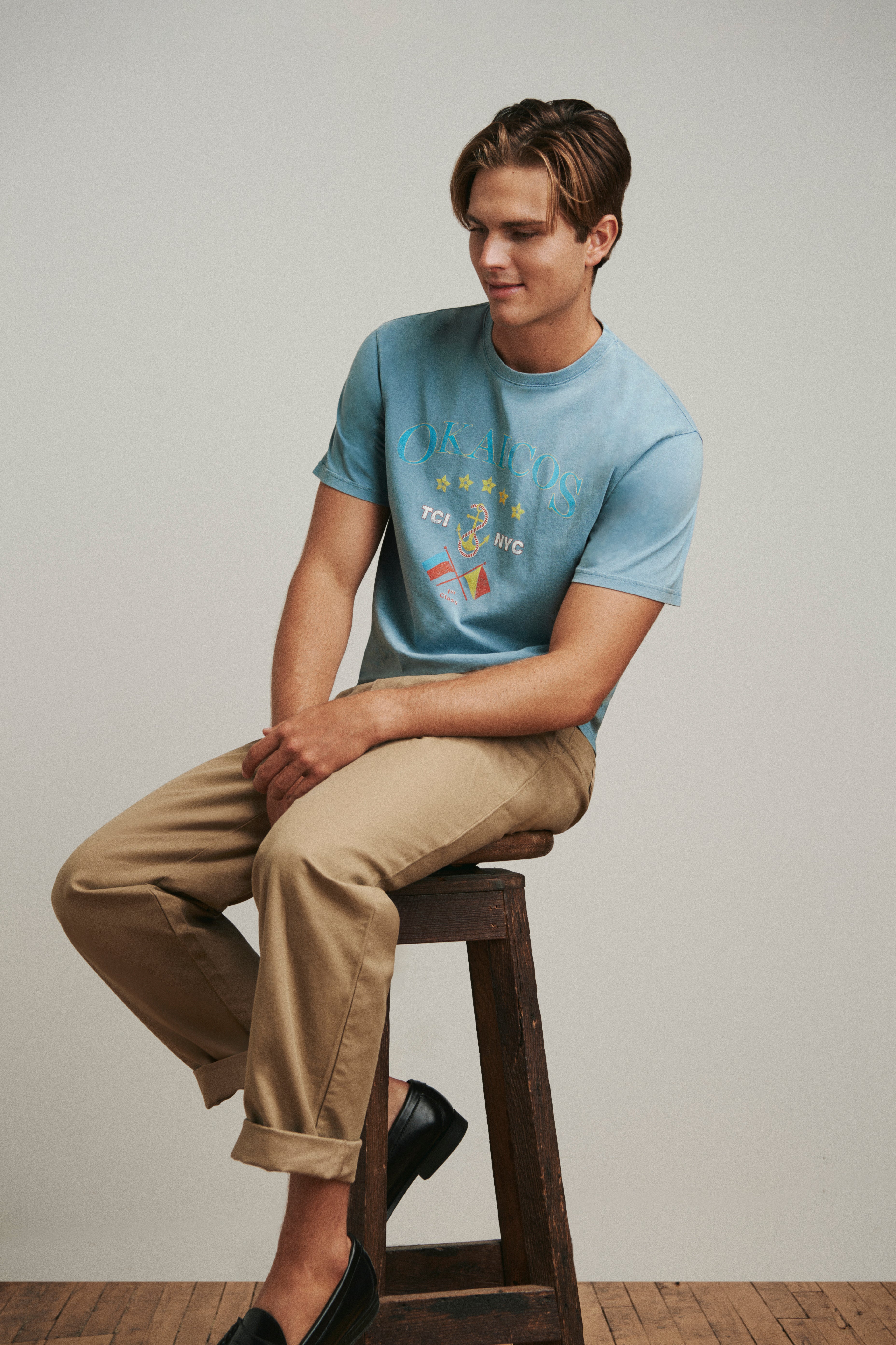 Man sitting on a wooden stool wearing a light blue t-shirt with graphics and beige pants.