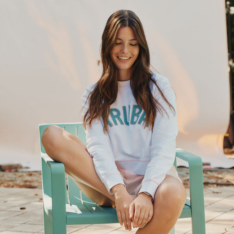 Woman sitting on a green chair wearing a white okaicos sweatshirt with 'ARUBa' text.
