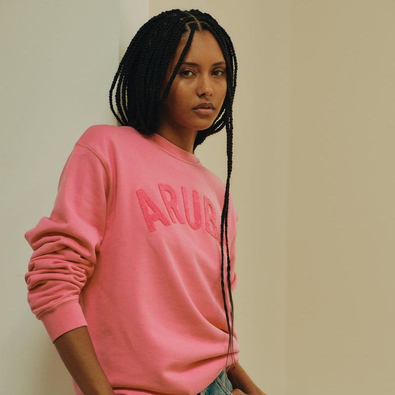 Person wearing a pink sweatshirt with 'ARUBA' text and denim shorts indoors.