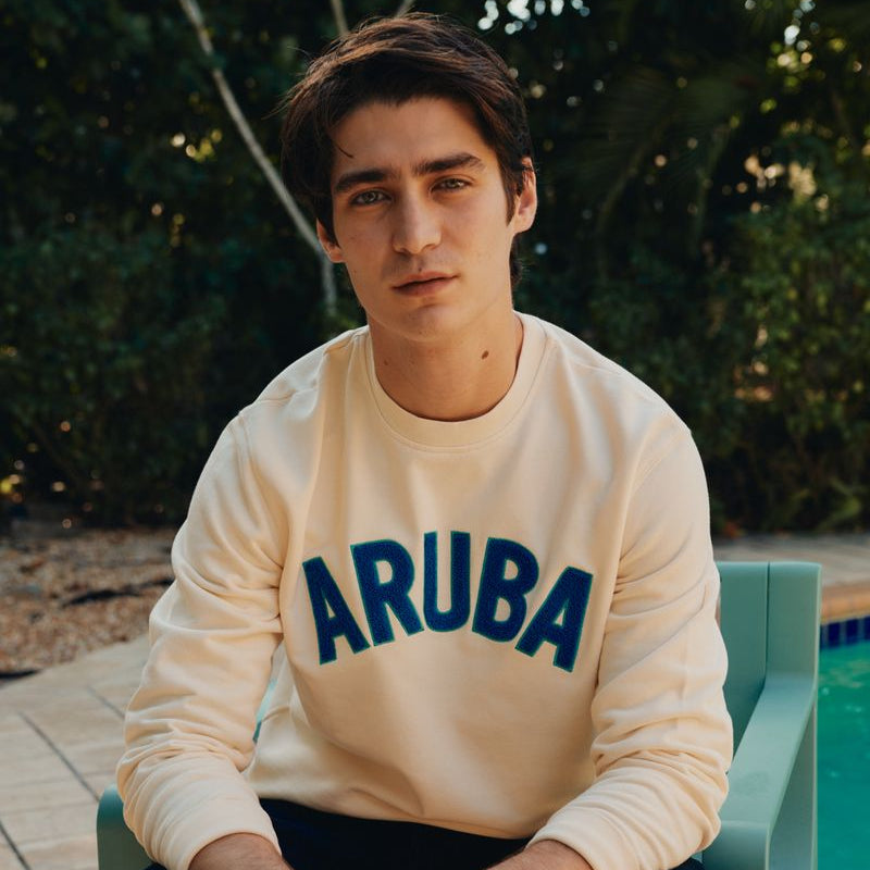 Person wearing a beige sweatshirt with 'ARUBA' printed on it, sitting outdoors.