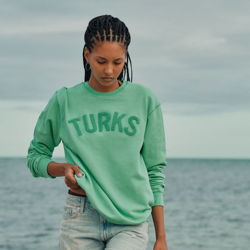 Person wearing a green sweatshirt with 'TURKS' text, standing by the ocean.