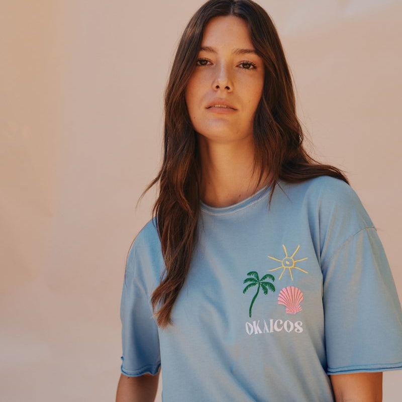 Woman wearing a light blue t-shirt with a palm tree and shell design on a plain background