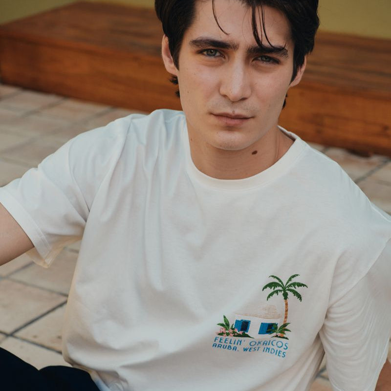 Person wearing a white t-shirt with a palm tree design on a tiled floor.