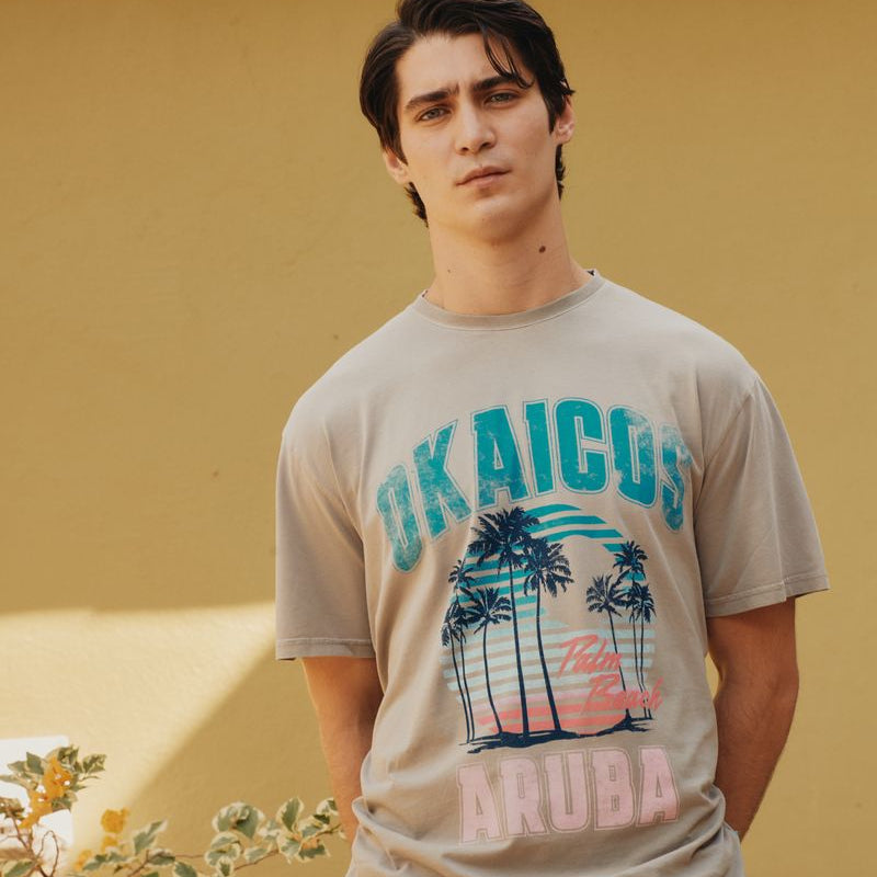 Man wearing a beige okaicos t-shirt with graphic design and text, standing against a yellow wall.