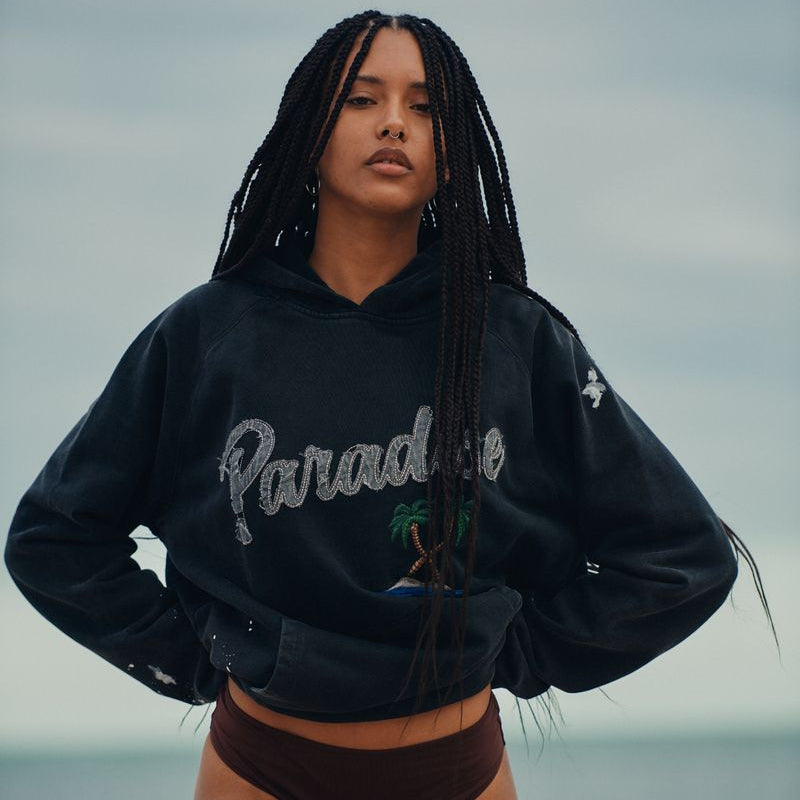 Woman wearing a black okaicos hoodie with 'Paradise' text and brown high-waisted shorts standing on a beach.