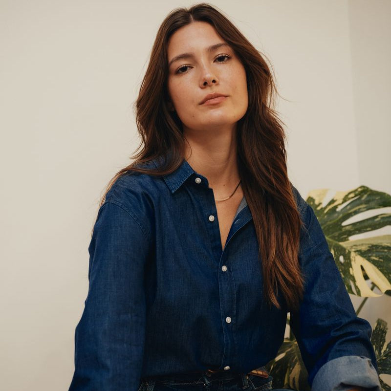 Woman wearing a blue denim shirt sitting on a wooden stool against a neutral wall with a plant in the background.
