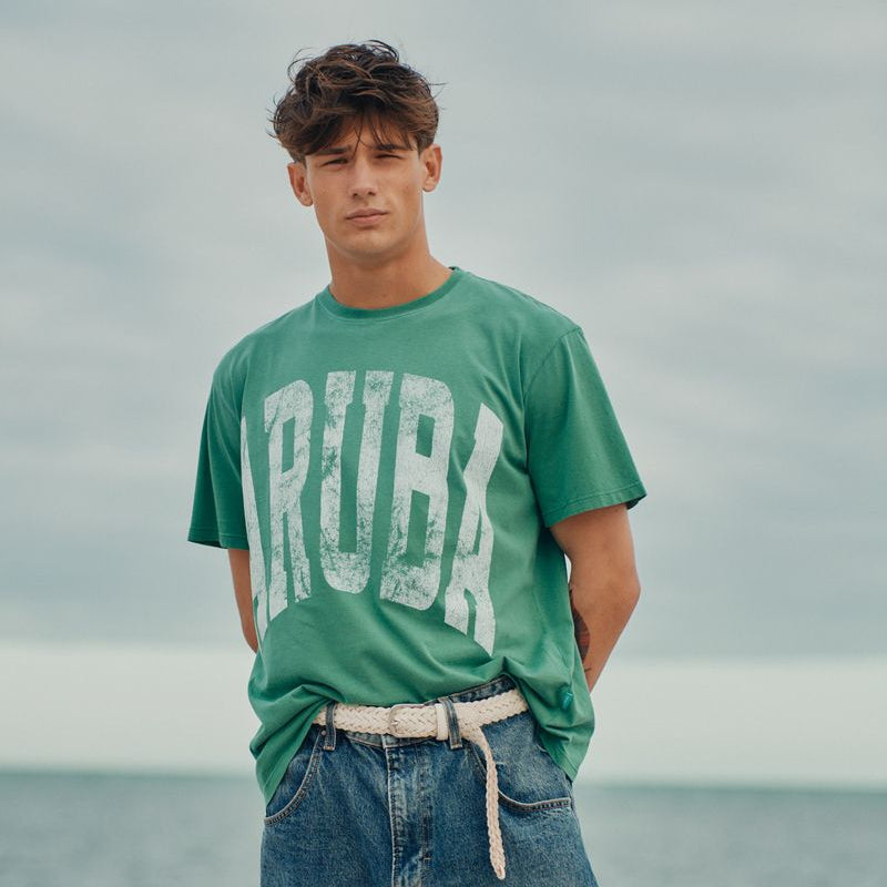 Man wearing a green Okaicos t-shirt with 'TRIBA' text on a beach
