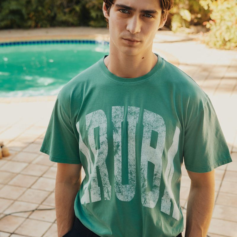 Person wearing a green Okaicos t-shirt with 'Aruba' printed on it, standing by a pool.