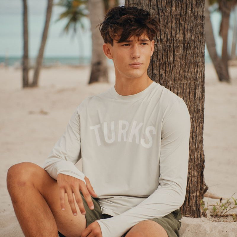 Person sitting on a sandy beach with palm trees in the background wearing an okaicos sun shirt with TURKS print
