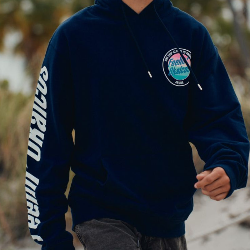 Person wearing a navy blue okaicos hoodie with a logo on a beach