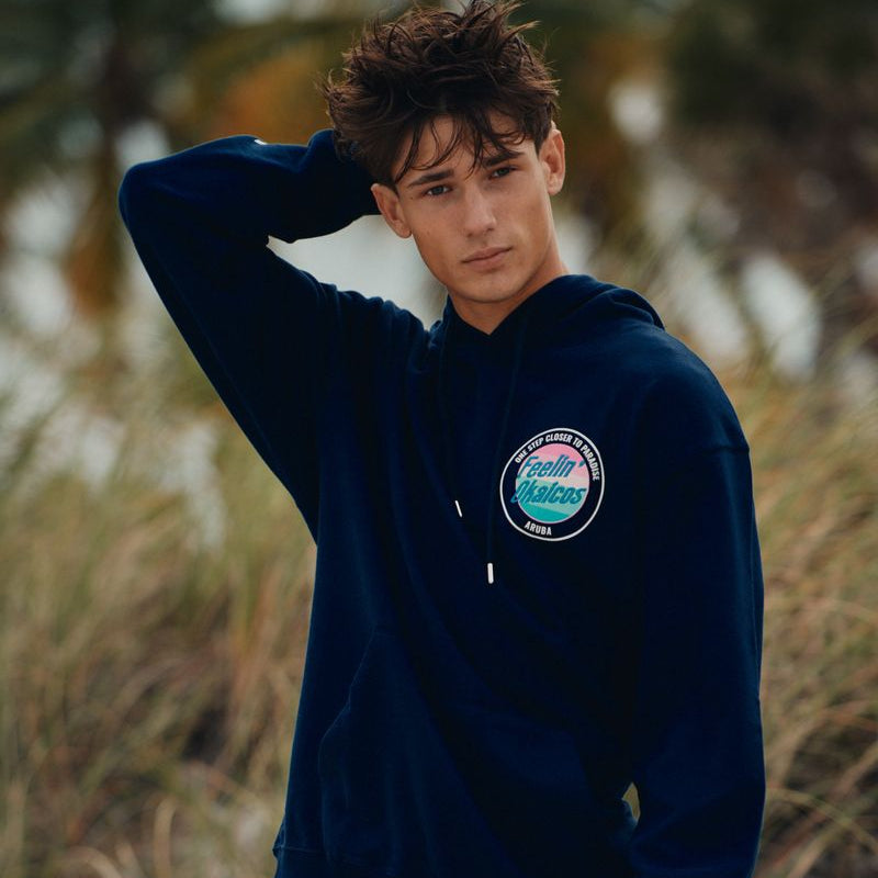 Person wearing a navy blue okaicos hoodie with a logo on a beach
