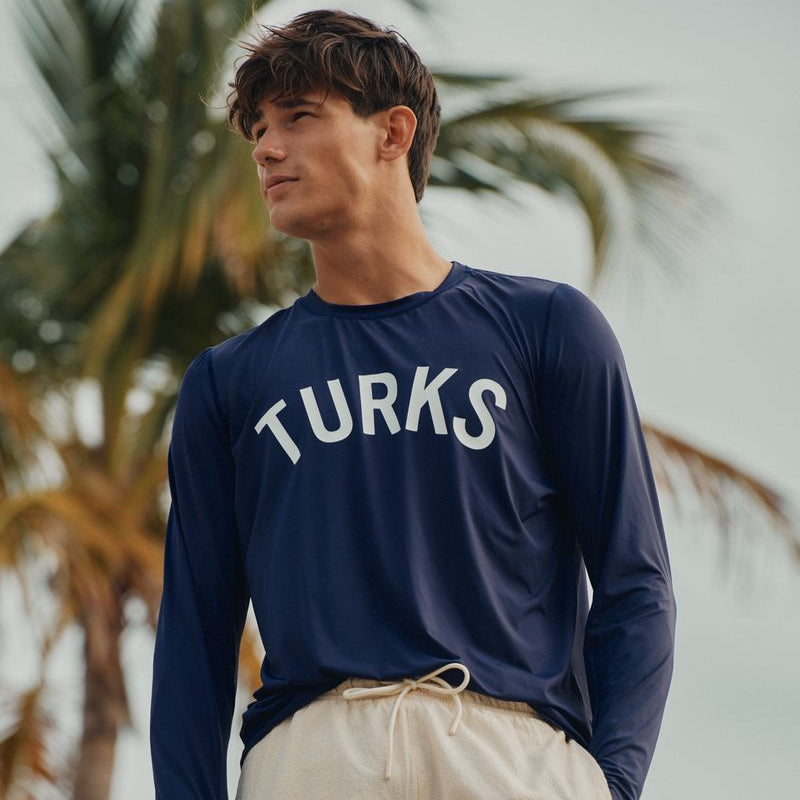 Man wearing a navy blue long-sleeve okaicos shirt with 'TURKS' text and beige shorts, standing in front of palm trees.