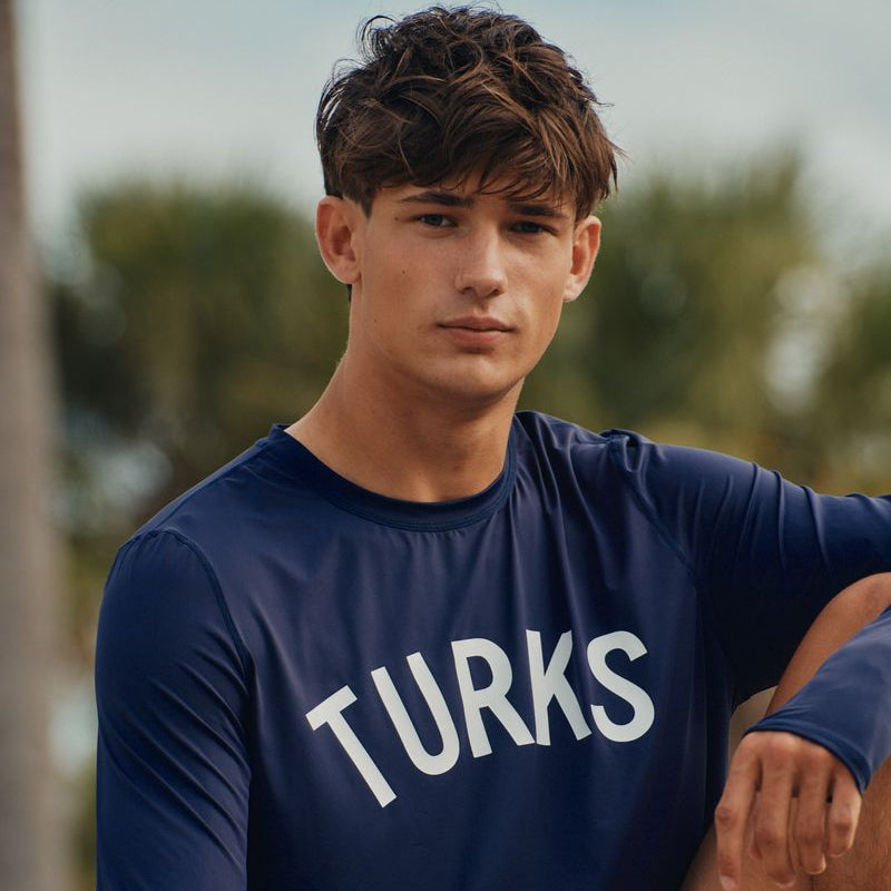 Man wearing a navy blue okaicos shirt with 'TURKS' text, sitting outdoors with blurred greenery in the background.