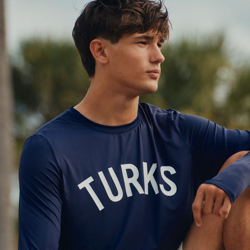 Person wearing a navy blue okaicos shirt with 'TURKS' text, sitting outdoors.