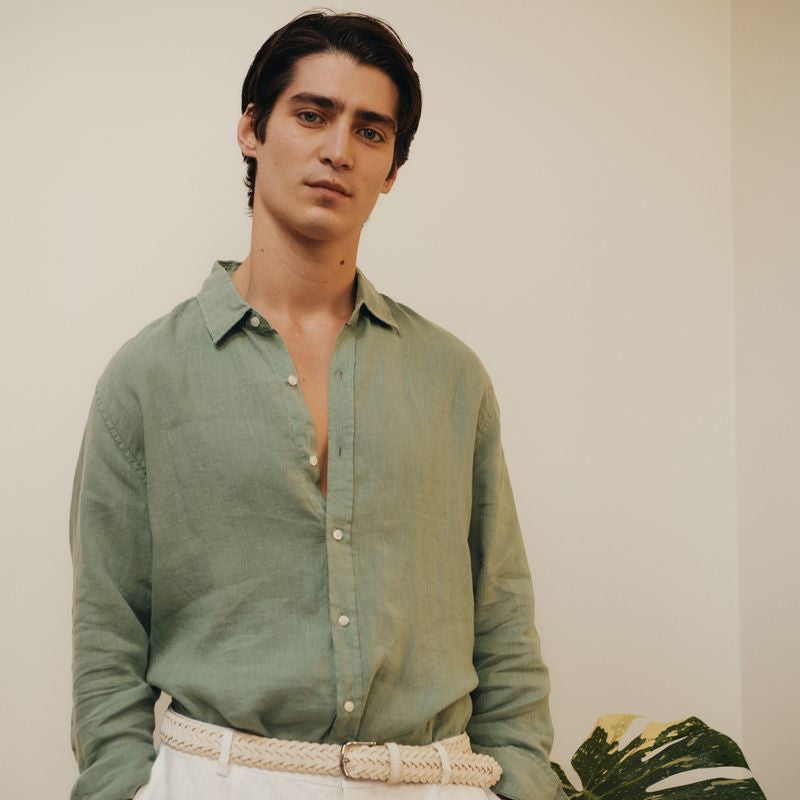 Man wearing a green okaicos shirt and white pants standing against a plain background with a plant.