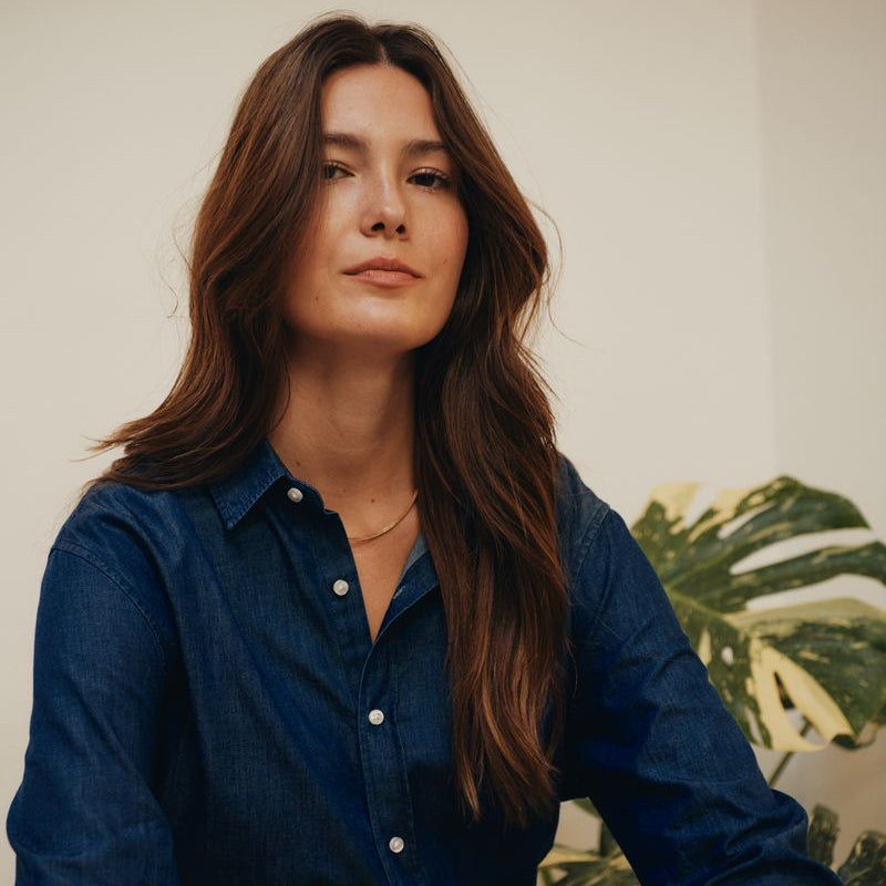 Woman wearing a blue denim shirt sitting in a room with a plant in the background