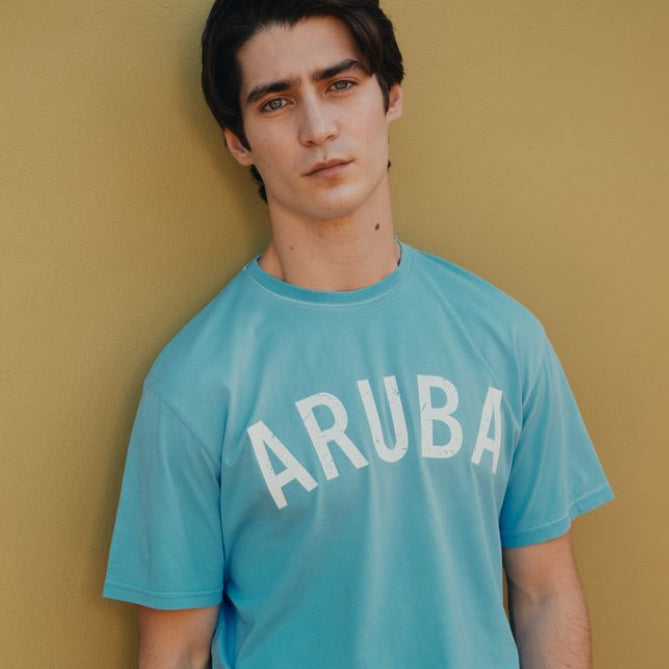 Man wearing a blue Okaicos 'ARUBA' t-shirt against a yellow wall.