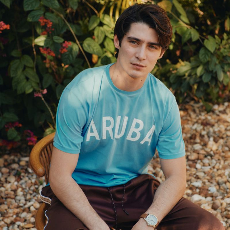 Man wearing a blue Okaicos 'ARUBA' t-shirt sitting outdoors with greenery in the background