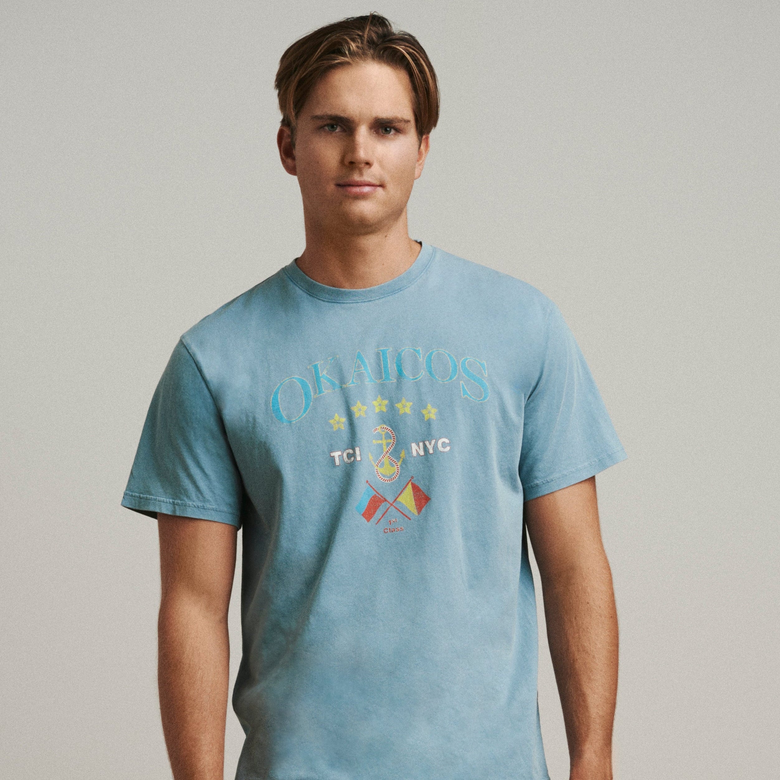 Man wearing a blue t-shirt with graphics and beige pants standing on a wooden floor.