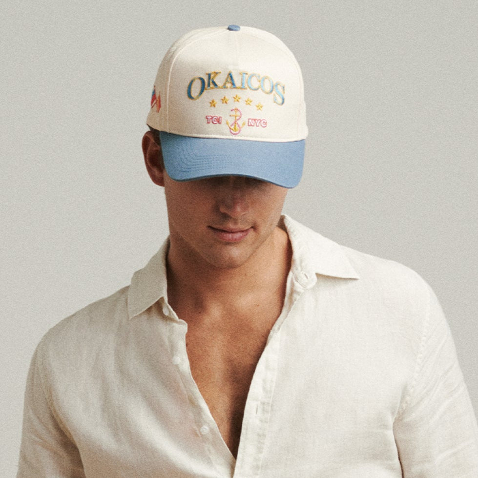 A person wearing a white shirt and an Okaicos souvenir hat with a cream crown and blue visor.