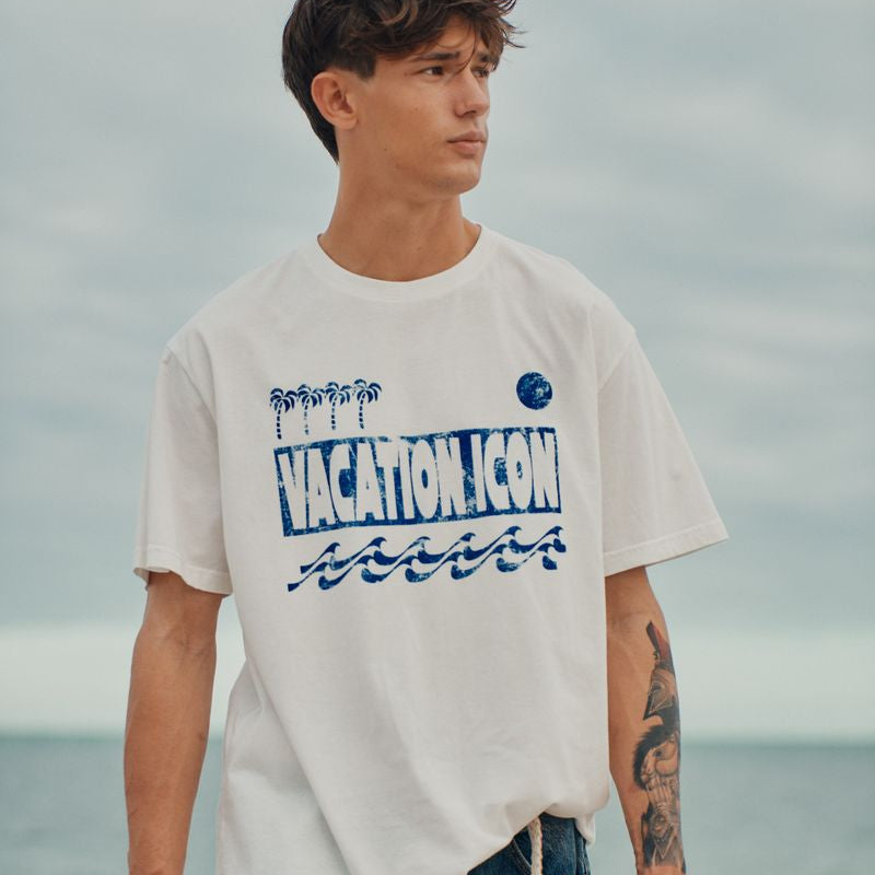 Man wearing a white okaicos t-shirt with blue vacation icon text and graphics, standing on a beach.