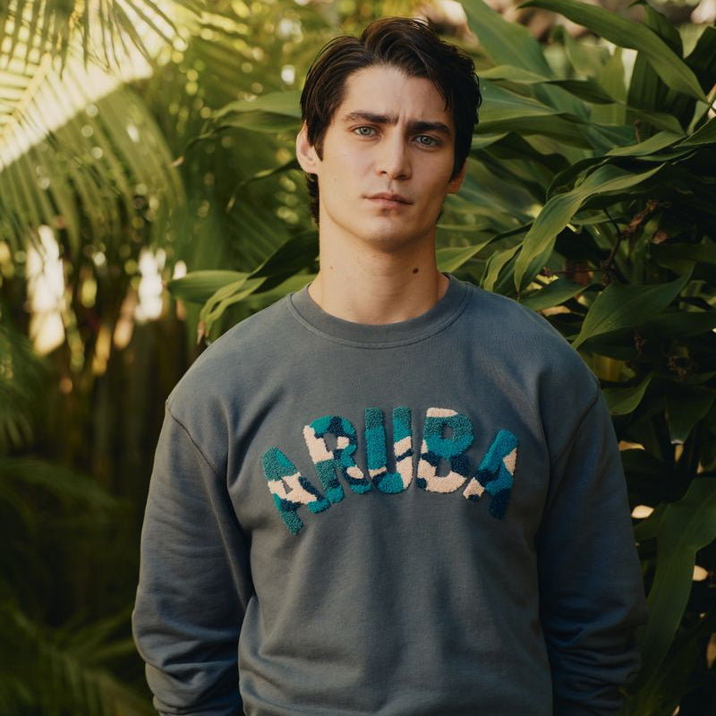 Man wearing a gray sweatshirt with colorful text standing in front of green foliage