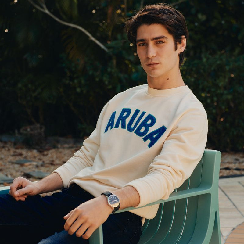 Man sitting on a green bench wearing a beige sweatshirt with 'ARUBA' text.