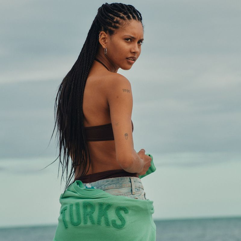 Woman with braided hair holding a green towel with 'Turks' text, standing on a beach.