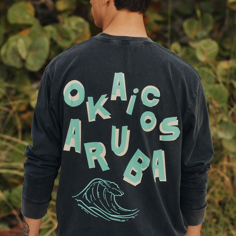 Person wearing a black sweatshirt with 'Okaic Aruba' text and wave design on the back, standing in front of green foliage.