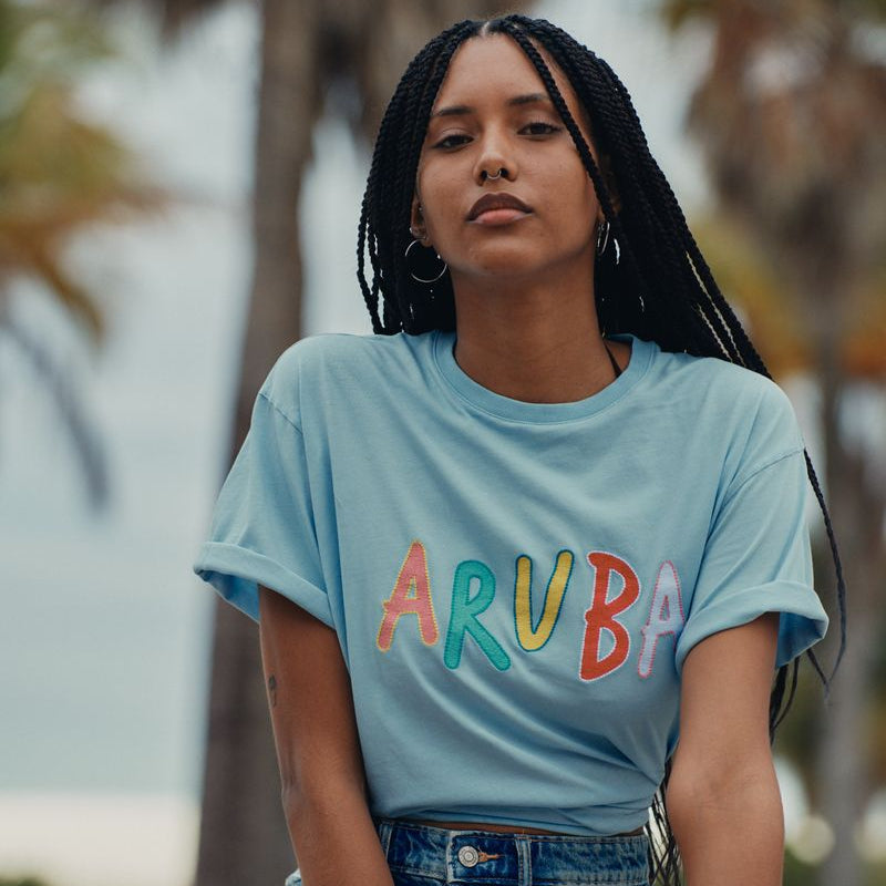 Woman wearing a light blue t-shirt with 'ARUBA' printed in colorful letters, sitting outdoors with blurred trees in the background.