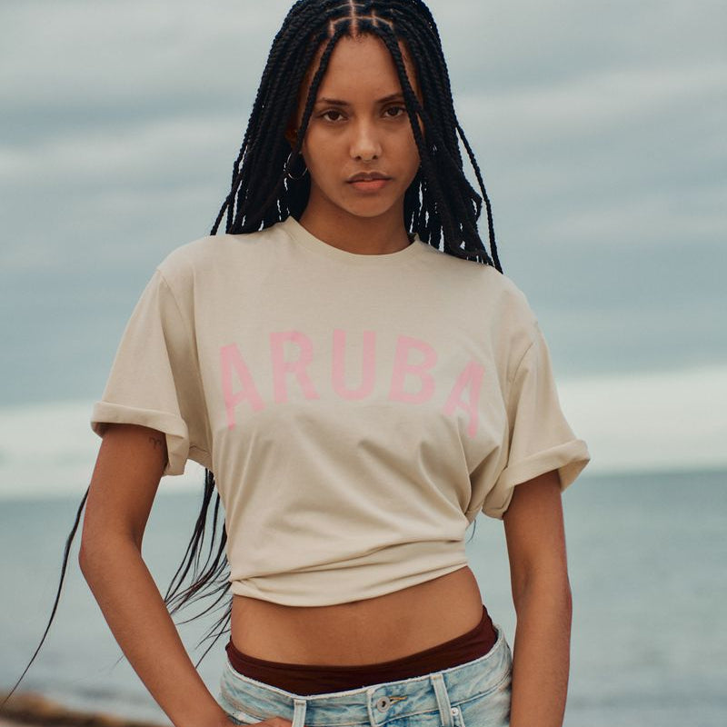 Woman wearing a beige t-shirt with 'ARUBA' printed on it, standing on a beach.